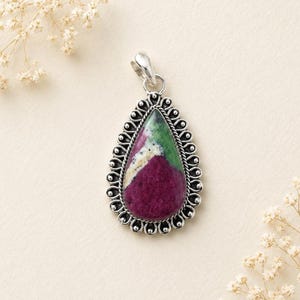 May include: A teardrop-shaped pendant featuring a multi-coloured stone with deep magenta, green, and white sections. The stone is set in a decorative sterling silver frame with a beaded edge and a small bail at the top for hanging on a chain.