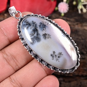May include: Oval pendant with a dendritic agate stone. The stone features a white and pale purple background with black and gray tree-like inclusions. The pendant has a silver-colored, ornate bezel and a loop for a chain.