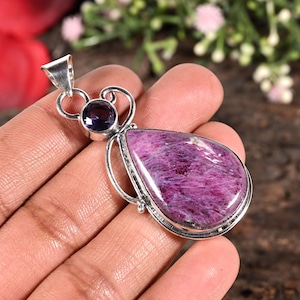 May include: A silver pendant with a large, teardrop-shaped, pinkish-purple gemstone. A smaller, round, dark purple stone is set above it. The pendant has a decorative silver bail and setting, highlighting the natural beauty of the stones.