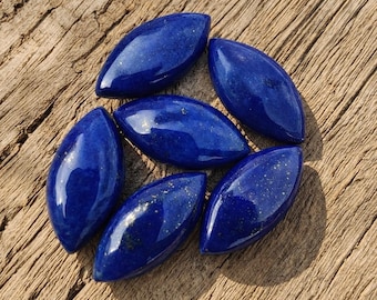 May include: Six polished marquise-shaped lapis lazuli gemstones with a deep blue color and subtle gold-colored flecks. Each stone features a smooth, rounded surface and a pointed oval silhouette, showcasing the natural mineral texture.
