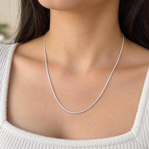 May include: A thin, silver-toned snake chain necklace with a smooth, rounded profile. The metallic chain rests against the neck, featuring a continuous, flexible design with a polished finish that reflects light along its entire length.