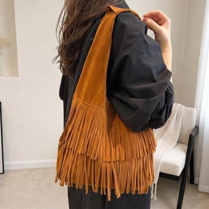 May include: A tobacco-coloured suede hobo bag featuring two horizontal tiers of long, dangling fringe that extend from the middle of the bag to the bottom edge, with a wide, integrated shoulder strap that rests comfortably over the shoulder.