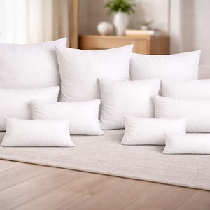 Puede incluir: Una colección de almohadas blancas de varios tamaños dispuestas sobre una alfombra de color neutro. Las almohadas son rectangulares y cuadradas, con un aspecto suave y esponjoso. La escena está bien iluminada, lo que sugiere un enfoque en la decoración del hogar y la comodidad.