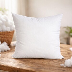 May include: A square, white pillow sits on a wooden surface, with fluffy white filling scattered around it. A wicker basket overflowing with more filling is in the background, suggesting a bedding or home decor theme.