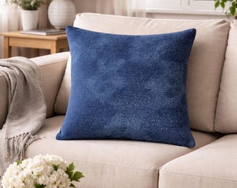May include: A square, navy blue decorative pillow with a subtle, textured pattern. The pillow is placed on a beige sofa in a well-lit living room setting. The pillow adds a touch of color and texture to the neutral-toned furniture.