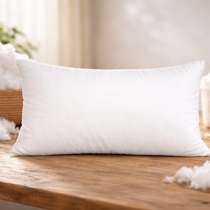 May include: A white rectangular pillow rests on a wooden surface, surrounded by fluffy white filling. A woven basket and a measuring cup filled with the same material are in the background. The pillow is the focal point, suggesting a bedding or home decor theme.