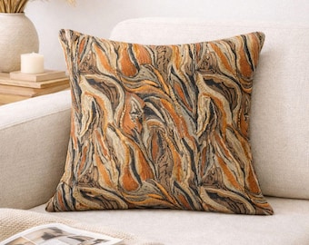 May include: Square decorative pillow with a woven pattern in shades of orange, brown, and black. The pillow is placed on a beige sofa, offering a warm and inviting aesthetic. The pillow's design features an abstract, organic motif.
