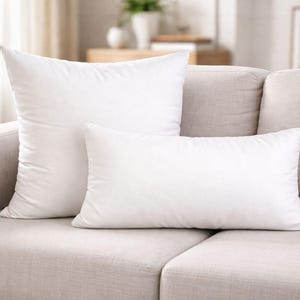 Puede incluir: Dos almohadas blancas, una rectangular y otra cuadrada, descansan sobre un sofá beige claro. Las almohadas son mullidas y parecen estar hechas de un material suave. El sofá está en una habitación bien iluminada.