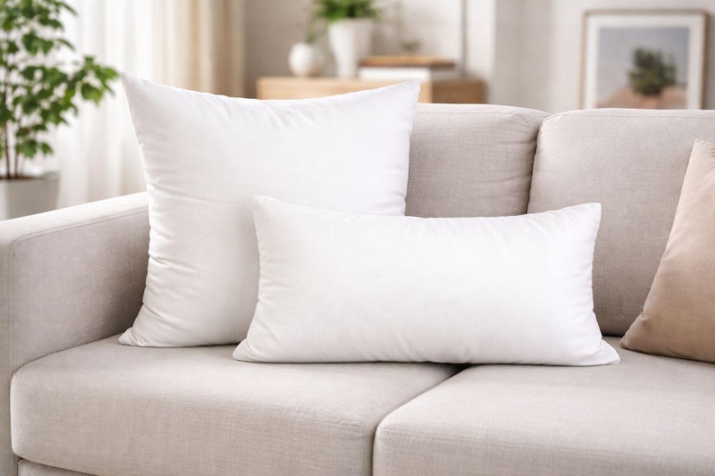 May include: Two white pillows, one rectangular and one square, rest on a light beige sofa. The pillows are fluffy and appear to be made of a soft material. The sofa is in a well-lit room.