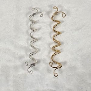 May include: Two spiral hair accessories. One is silver-colored, and the other is gold-colored. Both are made of wire and have a curly, wavy design. The ends of each accessory have a looped design.