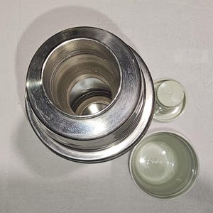 Może przedstawiać: Widok z g&oacute;ry na cylindryczny termos ze stali nierdzewnej z odsłoniętym metalowym otworem. Obok termosu znajduje się mały, jasnoszary plastikowy korek oraz oddzielny, pasujący jasnoszary kubek o gładkim wykończeniu wnętrza.