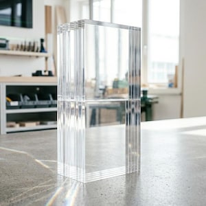 May include: A clear, rectangular acrylic block composed of multiple vertical panels. The transparent structure stands on a gray surface, reflecting light. The object is in a workshop setting, with tools and equipment visible in the background.