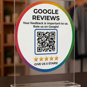 May include: A clear acrylic sign with a circular graphic promoting Google Reviews. The graphic features the words "GOOGLE REVIEWS", a QR code, and the text "GIVE US 5 STARS!". The sign is on a dark surface.
