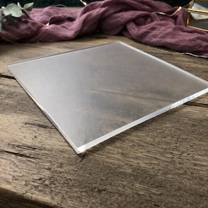 May include: A clear, square acrylic sheet with frosted edges. The sheet is positioned on a rustic wooden surface, with a soft, draped fabric in the background. The overall aesthetic is minimalist and clean, suitable for product photography or display.