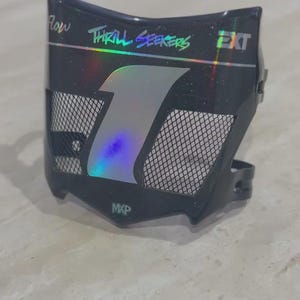 Peut inclure: Une plaque numéro de moto noire avec un grand chiffre un holographique au centre. La plaque comprend une grille d'aération, le texte "Flow", "THRILL SEEKERS", "EXT" et "MXP" en lettres holographiques, ainsi qu'une fine ligne horizontale holographique.