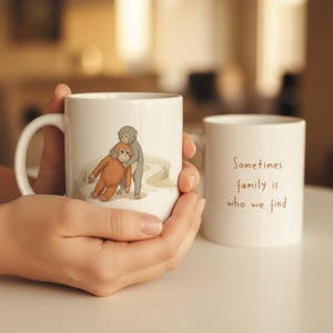May include: White ceramic mug featuring a cartoon illustration of two monkeys embracing. The second mug has the text "Sometimes family is who we find". The mugs are held in a person's hands.