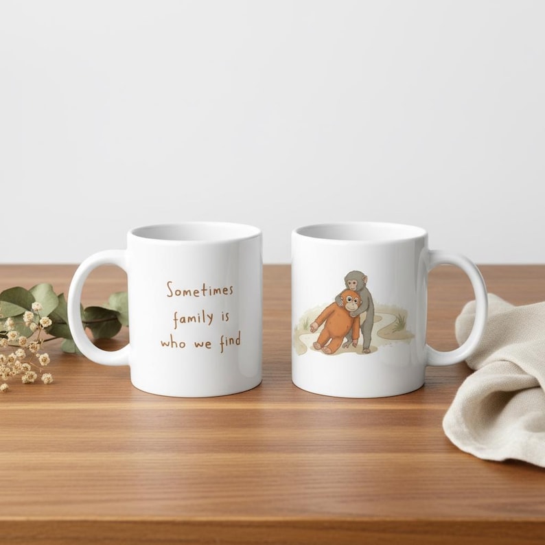 May include: Two white ceramic mugs with a brown handle are displayed on a wooden surface. One mug has the text "Sometimes family is who we find" in brown script. The other mug features an illustration of two monkeys hugging.