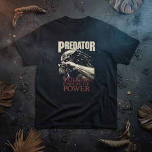 May include: Black t-shirt featuring a graphic of the Predator with the text "PREDATOR" above and "VULGAR DISPLAY OF POWER" below. The design is in shades of white and red.