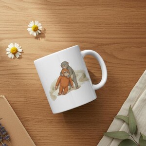 May include: White ceramic mug featuring a cartoon illustration of two orangutans. One orangutan is orange, and the other is gray. The mug has a curved handle and is set on a wooden surface with daisies.