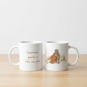 May include: Two white ceramic mugs. One mug has the text "Sometimes family is who we find" in brown. The other mug has an illustration of two monkeys hugging. The mugs are on a wooden surface.