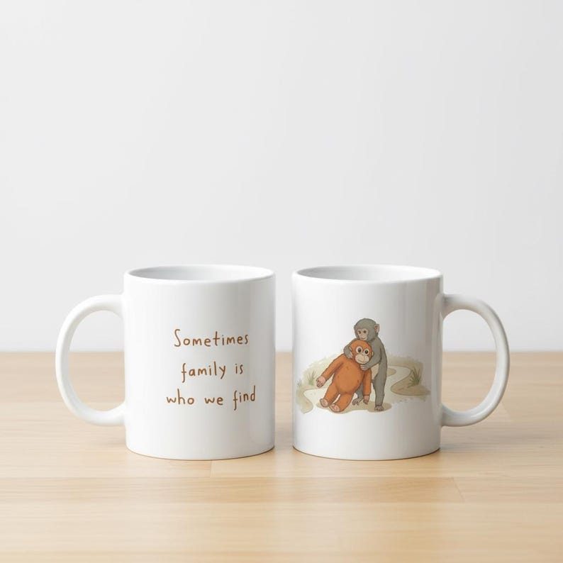 May include: Two white ceramic mugs. One mug has the text "Sometimes family is who we find" in brown. The other mug has an illustration of two monkeys hugging. The mugs are on a wooden surface.