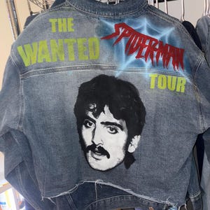 May include: A blue denim jacket featuring a black and white portrait of a person with a mustache on the back. The text THE WANTED is printed in lime green, SPIDERMAN in red with a blue web design, and TOUR in lime green. The hem is frayed.