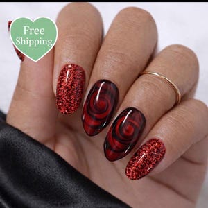 Puede incluir: Una mano muestra un conjunto de diseños de uñas rojas. Algunas uñas están cubiertas de purpurina roja, mientras que otras presentan un diseño de remolino en rojo y negro. Un anillo dorado adorna un dedo. Un corazón verde con la frase "Free Shipping" está en el fondo.