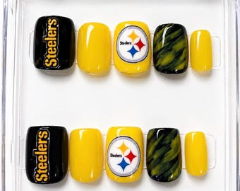 May include: A set of press-on nails in a clear plastic case. The nails feature a black base with "Steelers" in gold lettering, solid yellow, a yellow base with the Steelers logo, and a green and black marbled design.