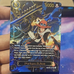 May include: A trading card featuring the character Portgas D. Ace from the Alabasta/Whitebeard Pirates series. The card has a blue and gold foil design with the number 5000 in the upper right corner. The card also includes game instructions.