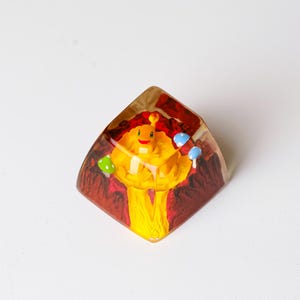 May include: A yellow and red resin keycap with a cartoon character design. The keycap features a smiling character with a crown, holding small objects. The background is a mix of red and brown, with small blue and green details.
