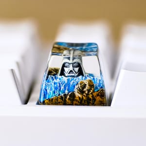 Puede incluir: Una tecla de teclado personalizada con el casco de Darth Vader dentro de resina transparente. El diseño incluye un paisaje azul y marrón, sobre un teclado blanco. Esta tecla única añade un toque personal a cualquier teclado mecánico.