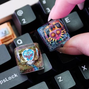 Könnte beinhalten: Zwei individuelle Tastenkappen mit Designs aus The Legend of Zelda. Eine Tastenkappe zeigt das Hylia-Schild, die andere Majora's Maske. Die Tastenkappen sind transparent mit farbenfrohen Kunstwerken, die auf einer schwarzen Tastatur liegen.