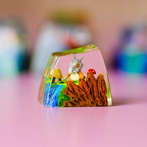May include: A clear resin keycap with a miniature landscape. It features a cartoon character with a skull helmet, a waterfall, mushrooms, and a red mushroom. The keycap is set against a pink background.