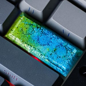 Puede incluir: Una tecla artesanal personalizada con un diseño abstracto vibrante. La tecla presenta una mezcla de tonos verdes, azules y amarillos, creando una apariencia texturizada única. La tecla es rectangular y se encuentra entre las teclas grises del teclado.