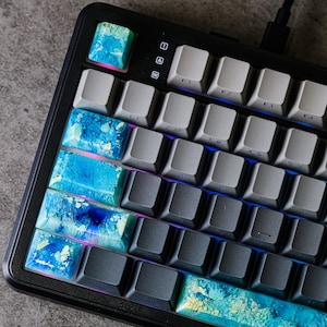 Puede incluir: Primer plano de un teclado mecánico negro con teclas personalizadas. Las teclas presentan un diseño veteado azul y verde. El teclado tiene teclas grises y negras, con un cable USB conectado.