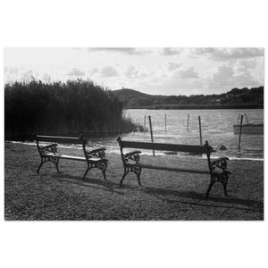 Puede incluir: Fotografía en blanco y negro de tres bancos de parque ornamentados junto a un lago. Los bancos tienen reposabrazos y respaldos decorativos. El lago tiene un pequeño bote y postes en el agua.