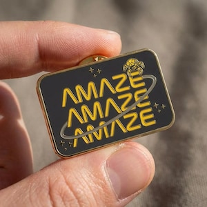 May include: A rectangular enamel pin with a gold border and pin back. The pin features the word "AMAZE" in yellow, stacked three times, with a stylized planet and stars on a black background. A small gold emblem is at the top.