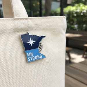 May include: A blue and white enamel pin in the shape of the state of Minnesota, with a starburst design and the text "MN STRONG". The pin is attached to a cream-colored tote bag.