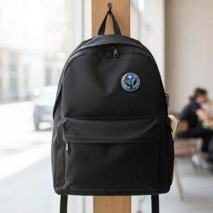 May include: Black backpack with a front pocket and a small round emblem. The emblem features a bird design with a star and a gradient of blue, purple, and green colors. The backpack is hanging on a wooden hook.
