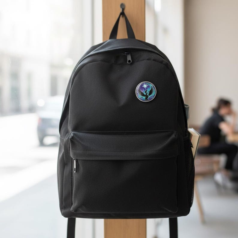 May include: Black backpack with a front pocket and a small round emblem. The emblem features a bird design with a star and a gradient of blue, purple, and green colors. The backpack is hanging on a wooden hook.