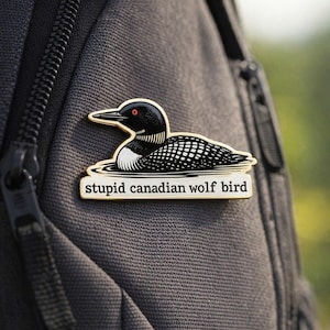 May include: A black, white, and gold enamel pin featuring a loon design. The pin has the text "stupid canadian wolf bird" on a white banner. The pin is attached to a gray backpack.