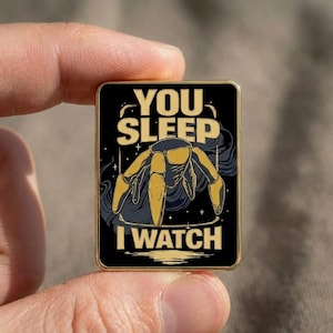May include: A rectangular pin with a black background and gold trim. The pin features a stylized yellow spider with the text "YOU SLEEP I WATCH" in gold lettering. The design includes a dark blue smoky background and small stars.