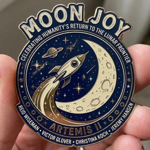 May include: A circular, navy blue and gold pin with the words "MOON JOY" and "CELEBRATING HUMANITY'S RETURN TO THE LUNAR FRONTIER". The pin features a rocket ship, moon, and stars, with the text "ARTEMIS II" and astronaut names.