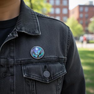 May include: A round, silver-toned pin featuring a stylized bird design with a star. The bird has teal and purple wings against a background of swirling colors. The pin is attached to a dark gray denim jacket.