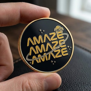May include: A round, black coin with gold lettering that reads "AMAZE" three times, with a gold planet-like ring around the text. The coin has a gold rim and a small gold graphic of a creature. The coin is held in a hand.