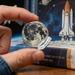 May include: A round, silver-toned pin featuring a detailed depiction of the moon's surface. The pin has a small, white banner with the text "Copy, Moon joy". The pin is held in front of a space-themed background.