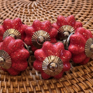 May include: A set of red ceramic cabinet knobs with a crackle effect. Each knob features a flower design, a gold-coloured metal base, and a silver-coloured centre. The knobs are displayed on a woven surface.