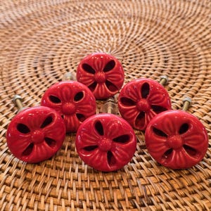 May include: Six red ceramic cabinet knobs with a floral design. Each knob has a glossy finish and is mounted on a metal screw. The knobs are arranged on a woven, light brown surface.
