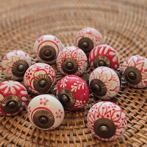 May include: A collection of decorative ceramic cabinet knobs. The knobs are white and red with floral patterns and brass hardware. The knobs are arranged on a woven surface. The knobs are approximately 4 cm in diameter.