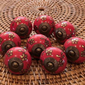 May include: Eight red ceramic cabinet knobs with floral designs in gold and black. Each knob has a bronze-coloured base and a rounded shape. The knobs are arranged on a woven, light brown surface.